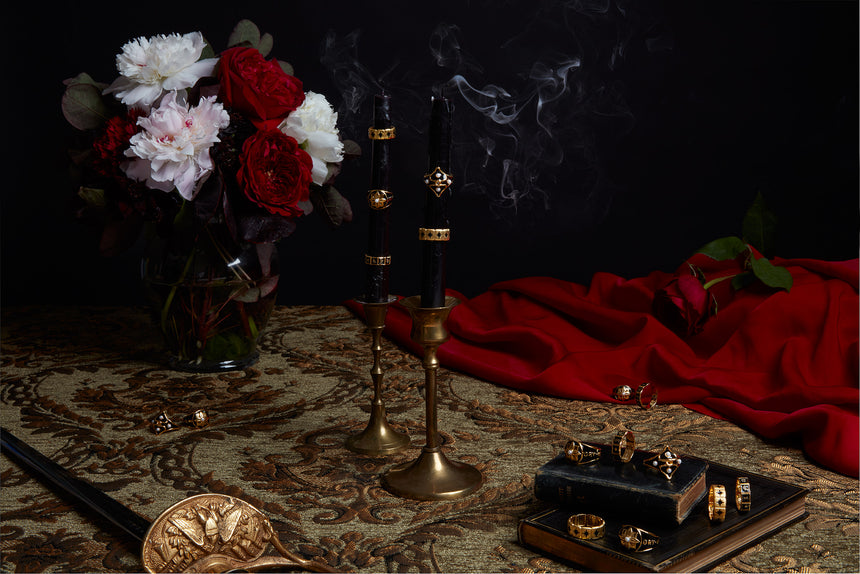Antique inspired mourning rings with black enamel, sitting on antique books and black candles that have been blown out, on beautiful antique candle holders. Fresh flowers and tapestries complete the image.